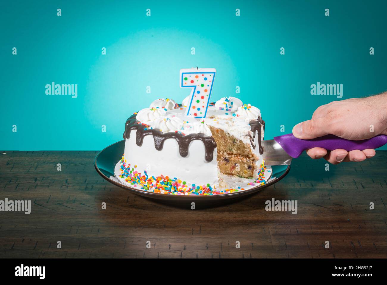 A birthday cake, missing a slice, bears a candle in the shape of the number 7 while a hand cuts another slice. Stock Photo