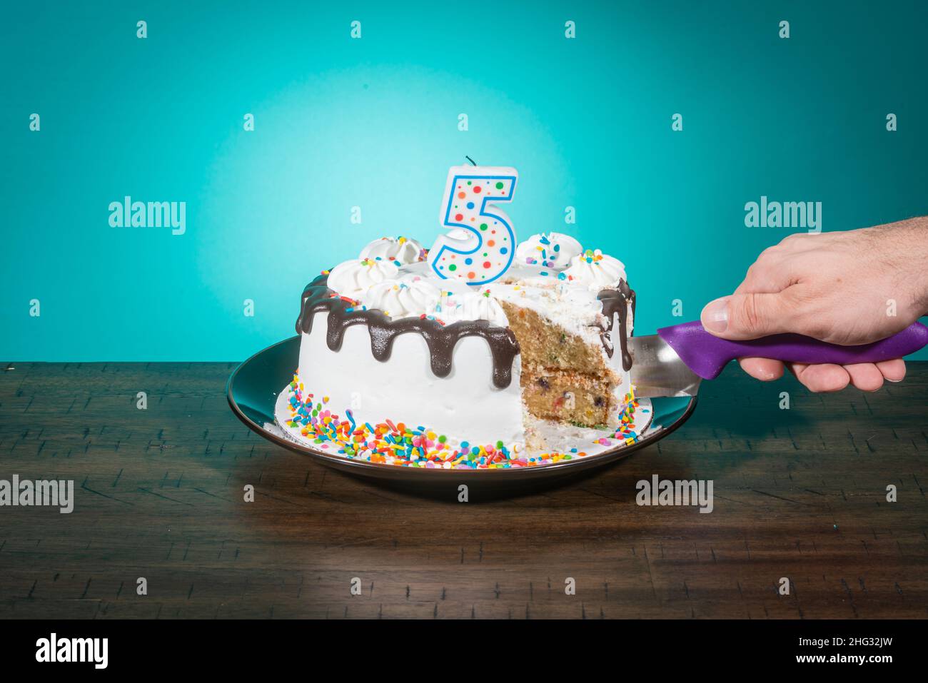 A birthday cake, missing a slice, bears a candle in the shape of the number 5 while a hand cuts another slice. Stock Photo