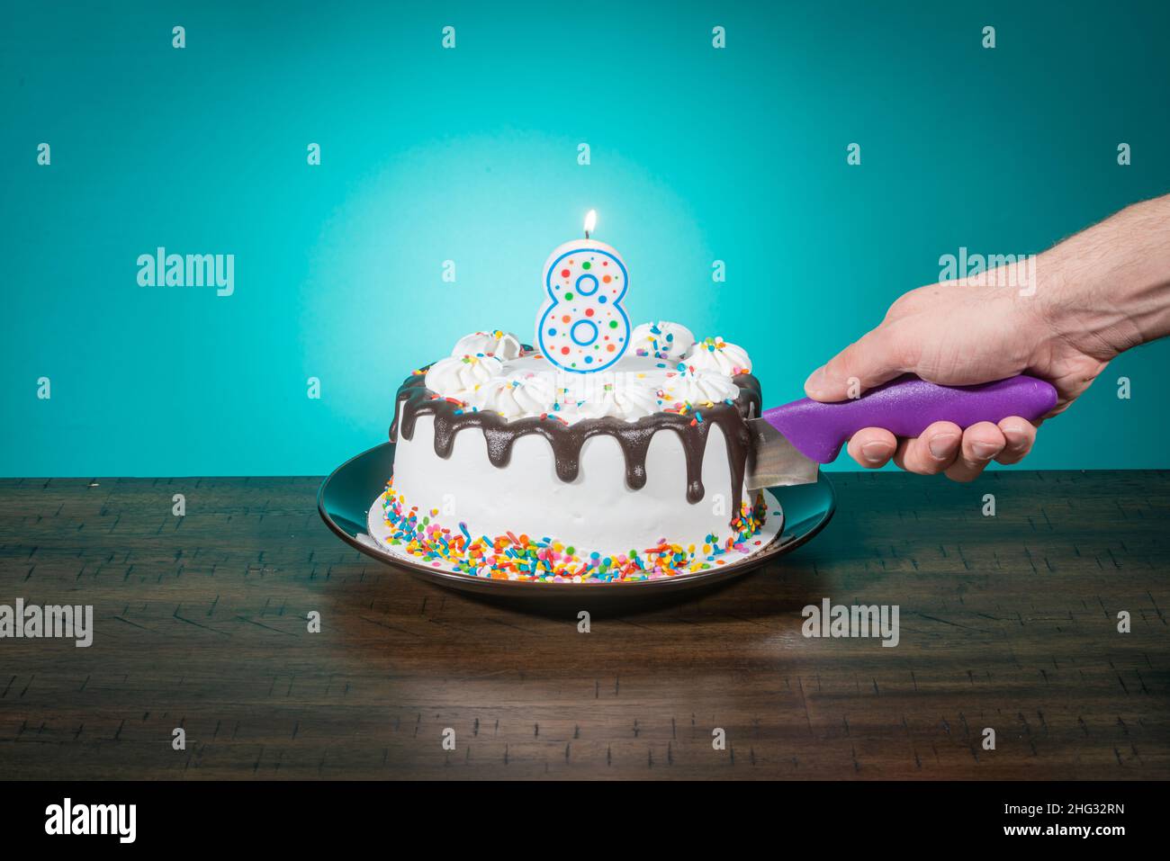 A birthday cake bears a candle in the shape of the number 8 while a hand cuts a slice. Stock Photo