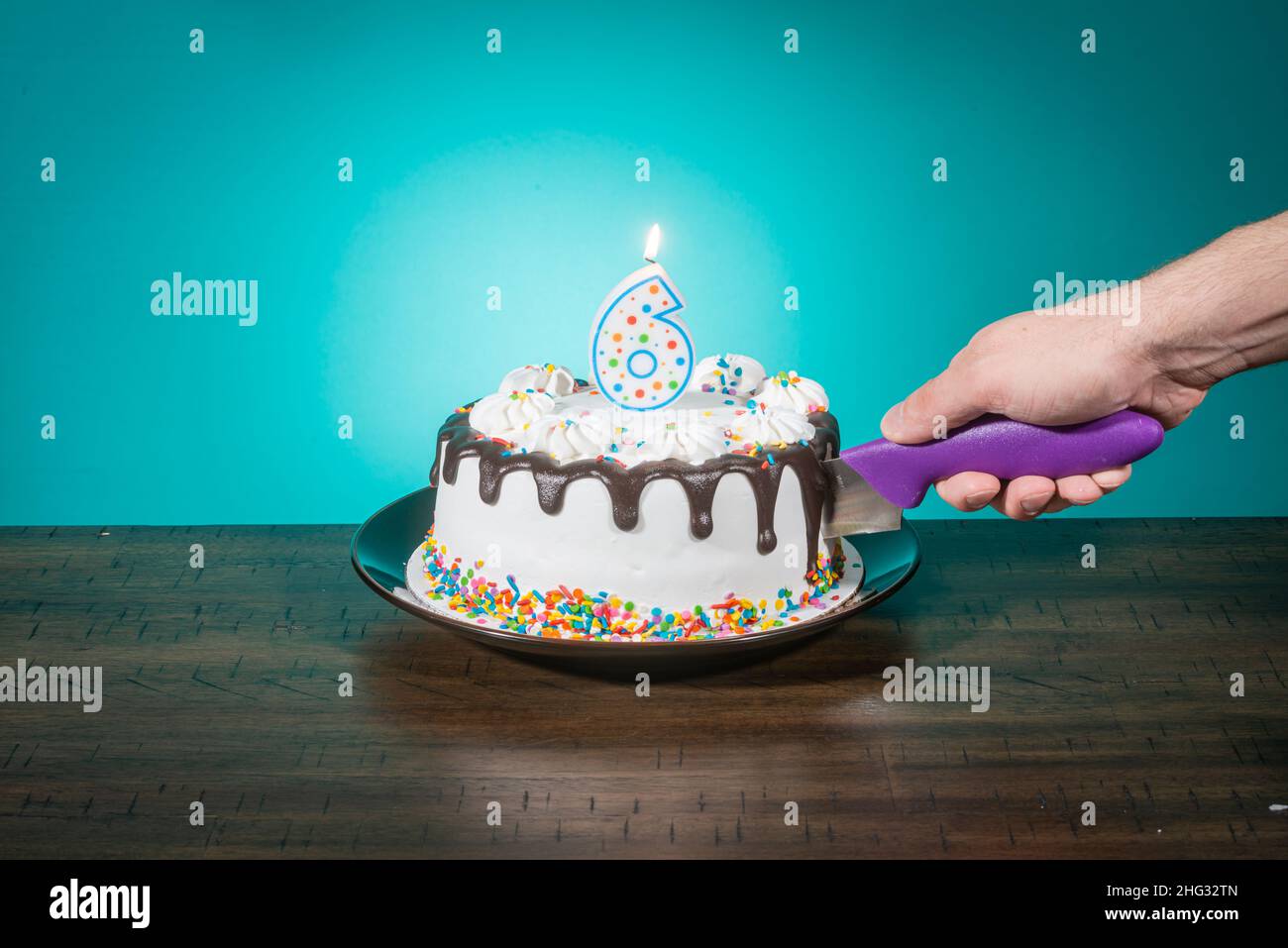 A birthday cake bears a candle in the shape of the number 6 while a hand cuts a slice. Stock Photo