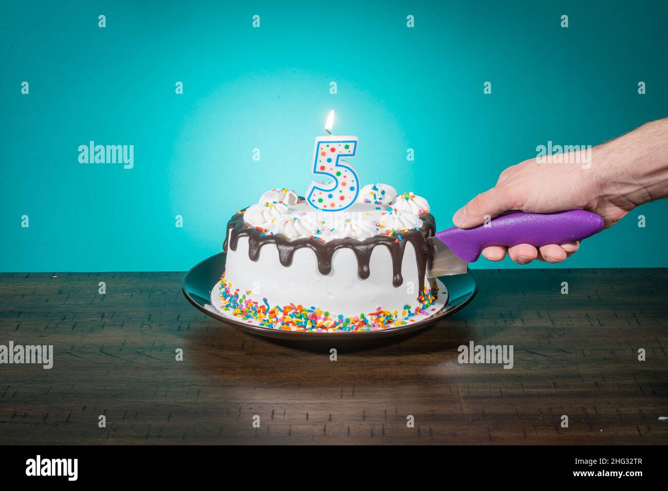 A birthday cake bears a candle in the shape of the number 5 while a hand cuts a slice. Stock Photo