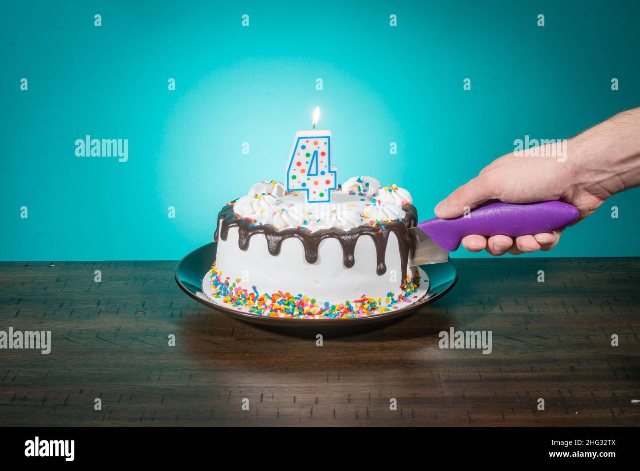 A birthday cake bears a candle in the shape of the number 4 while a hand cuts a slice. Stock Photo