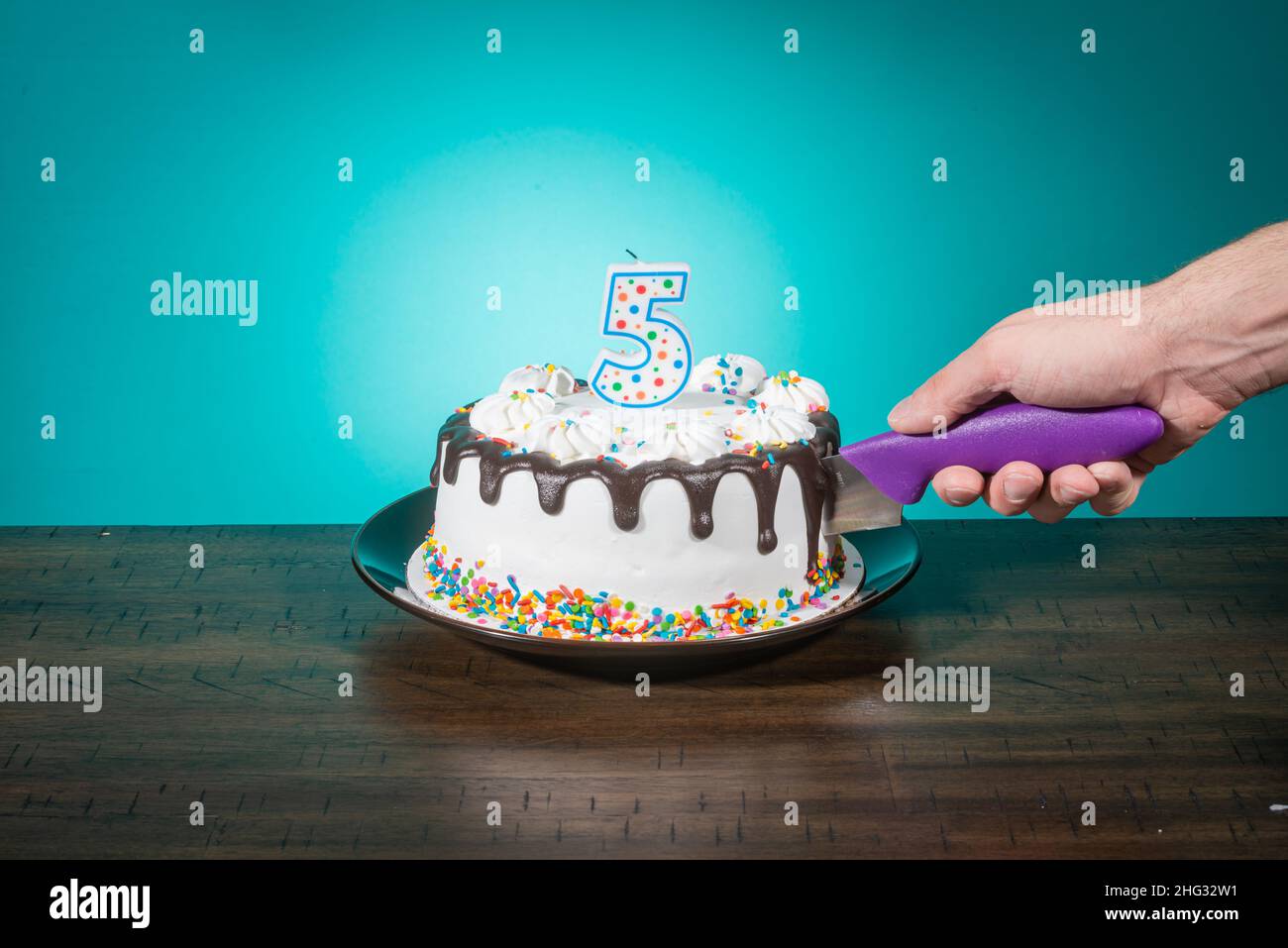 A birthday cake bears a candle in the shape of the number 5 while a hand cuts a slice. Stock Photo