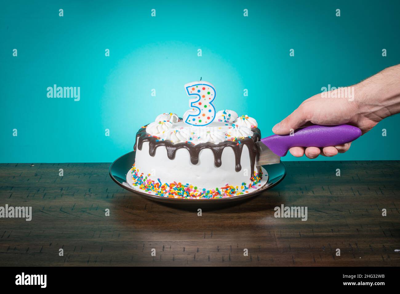 A birthday cake bears a candle in the shape of the number 3 while a hand cuts a slice. Stock Photo
