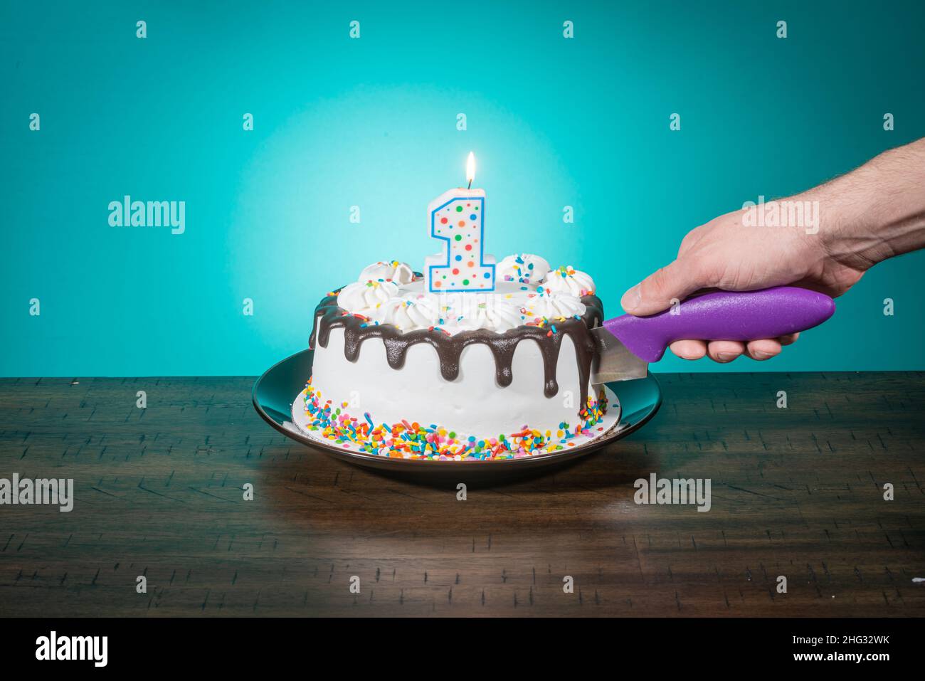A birthday cake bears a candle in the shape of the number 1 while a hand cuts a slice. Stock Photo