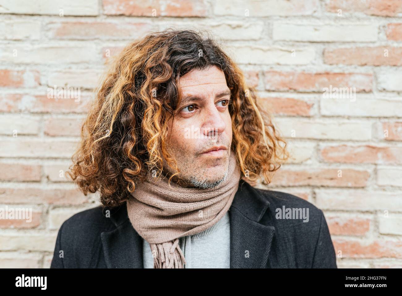 Mature man in scarf with curly hair looking away while standing against brick wall in city Stock Photo