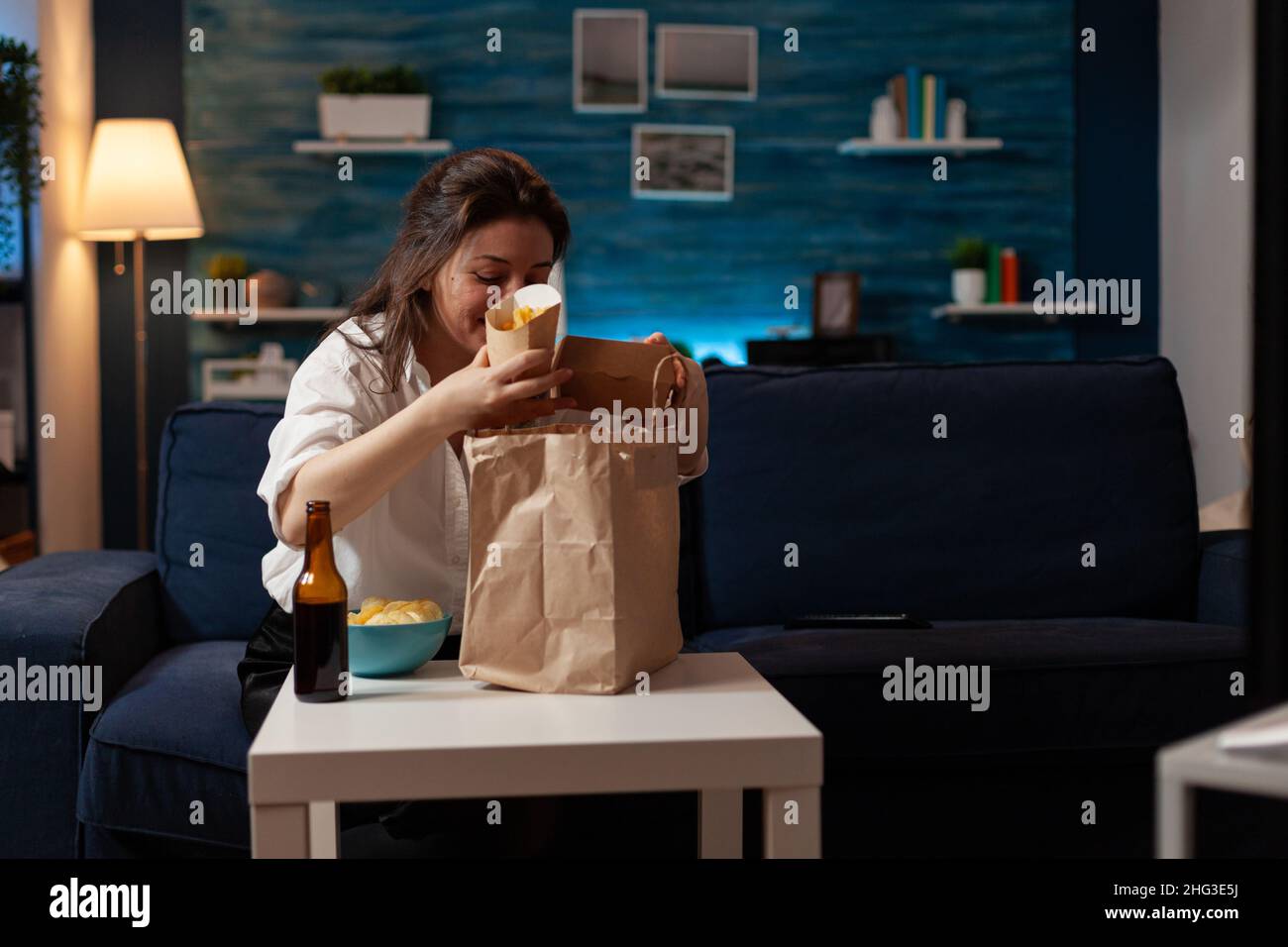 Woman in living room preparing for couch dinner unpacking french fries and tasty hamburger from takeaway paper bag. Person having burger junk food meal and bottled beer sitting on sofa. Stock Photo