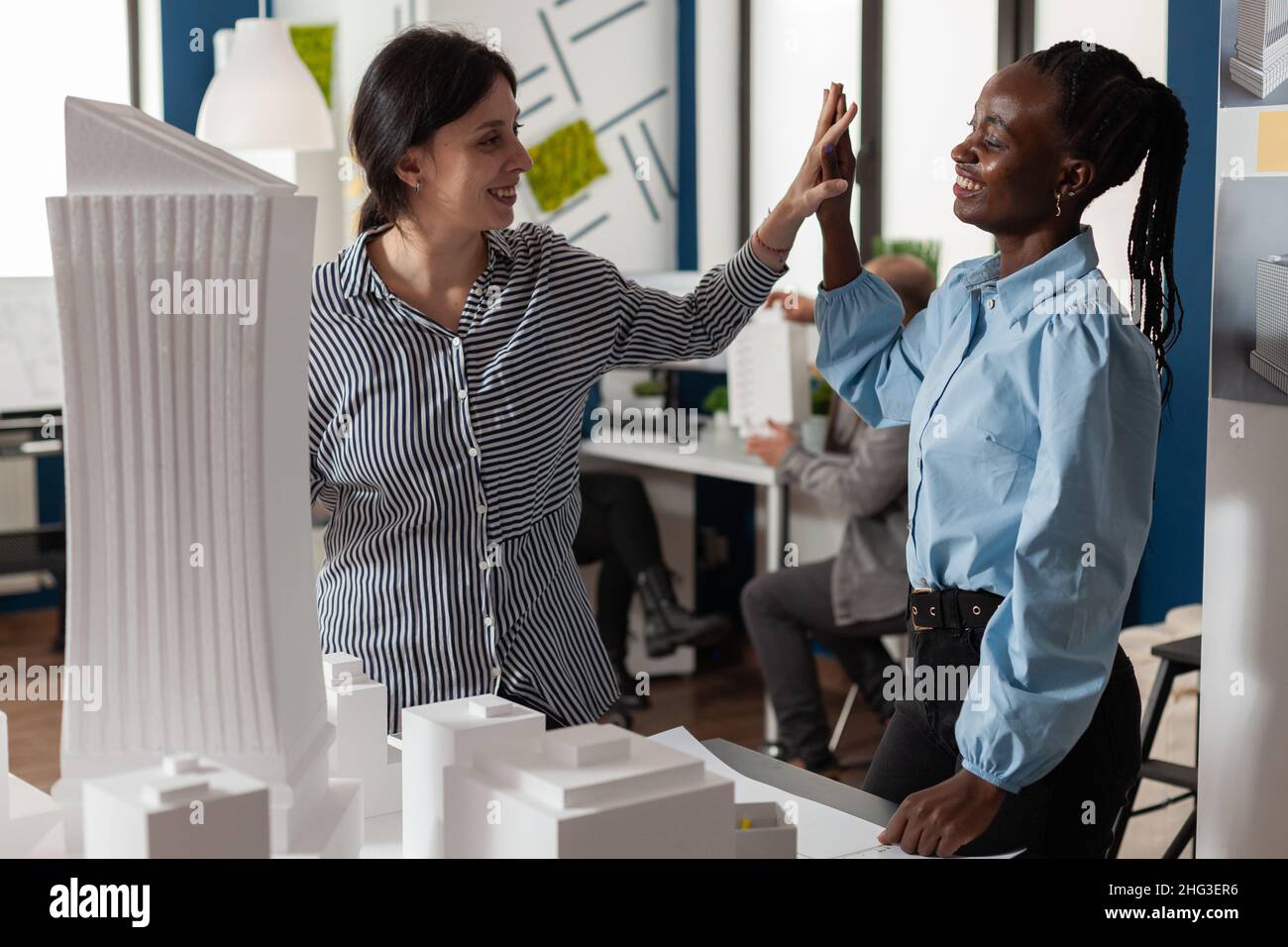 Team of two women architects high fiving standing in front of table with foam scale model of residential buildings in architectural modern office. Successful happy engineers celebrating team project. Stock Photo