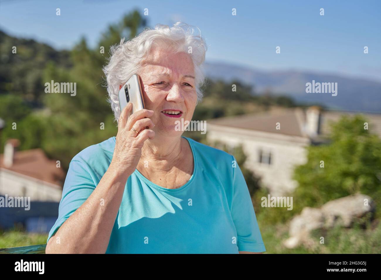 Positive elderly female with gray hair having conversation on mobile phone in sunny day against green mountains with cottages Stock Photo