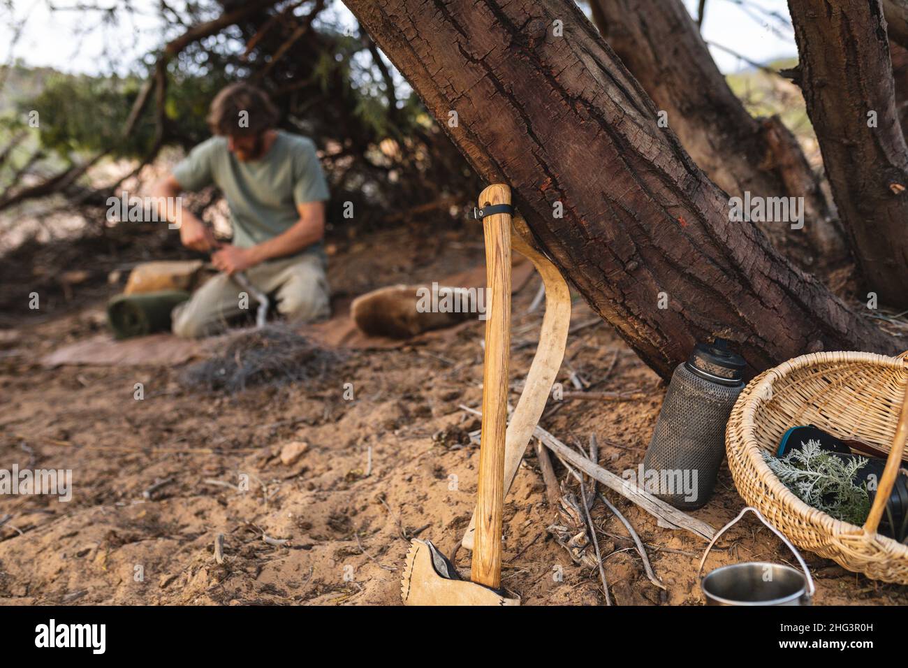 Axe leaning on tree trunk in front of male caucasian survivalist kneeling in forest Stock Photo
