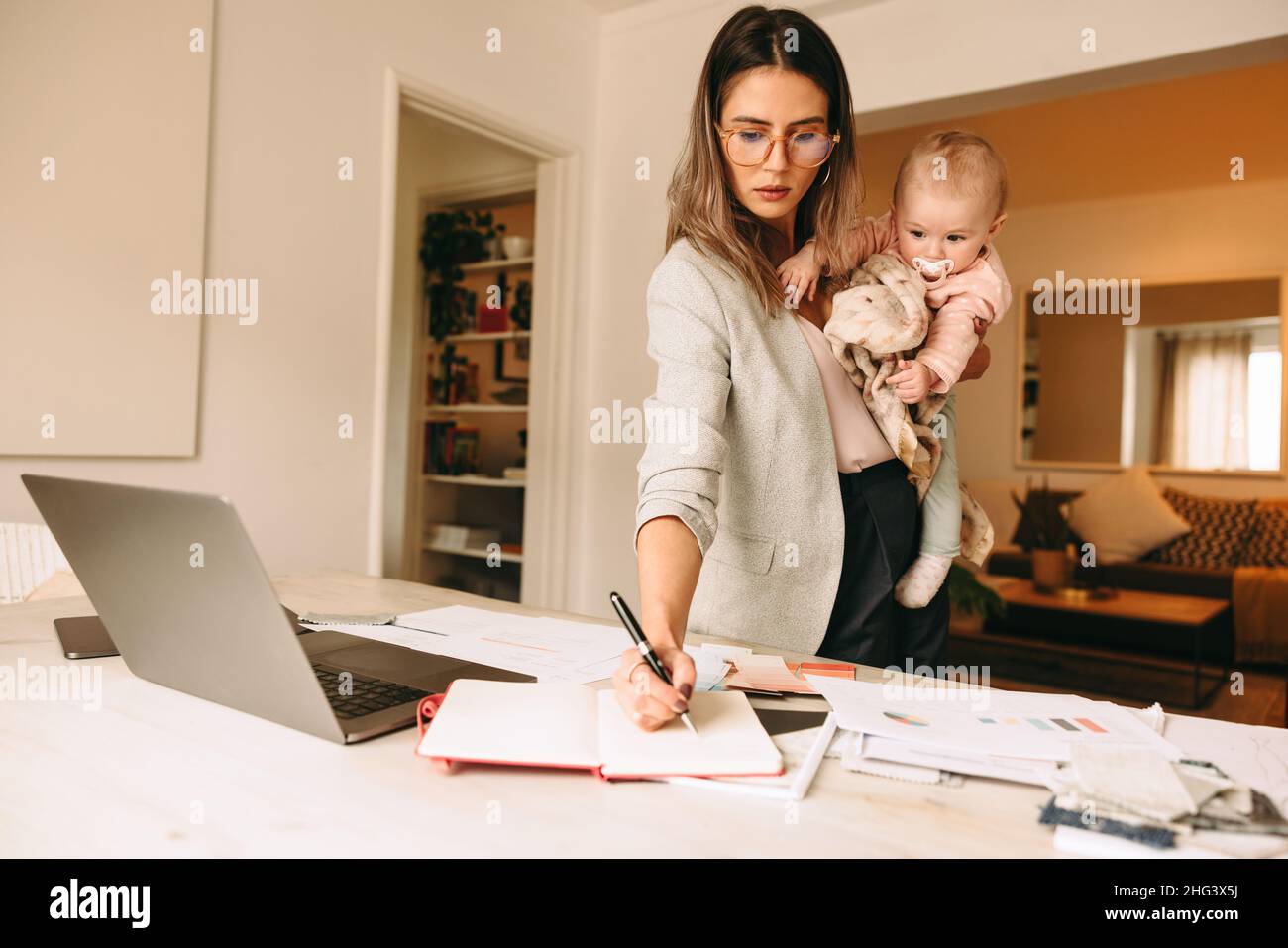 Female interior designer making notes while holding her baby. Multitasking mom planning a new project in her home office. Creative businesswoman balan Stock Photo