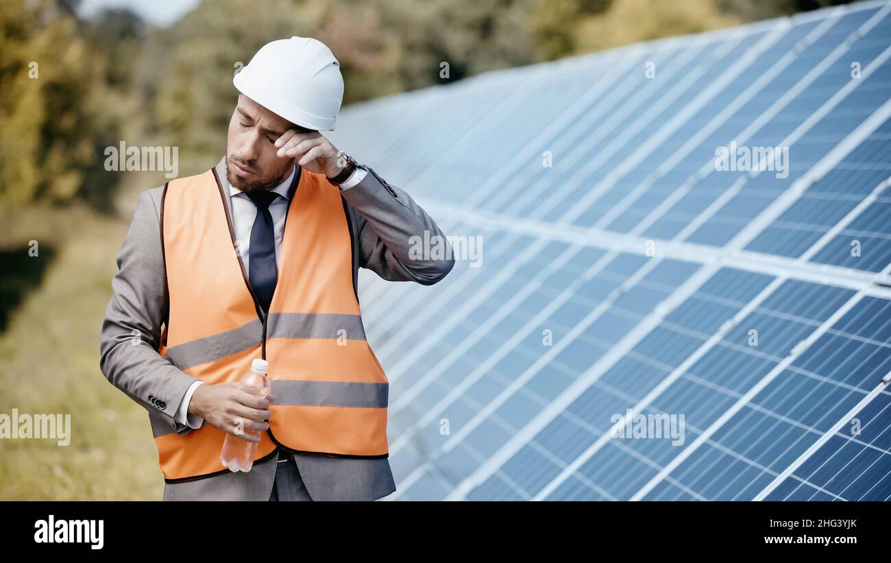businessman in safety vest rubbing eye and holding bottle with water near solar panels Stock Photo
