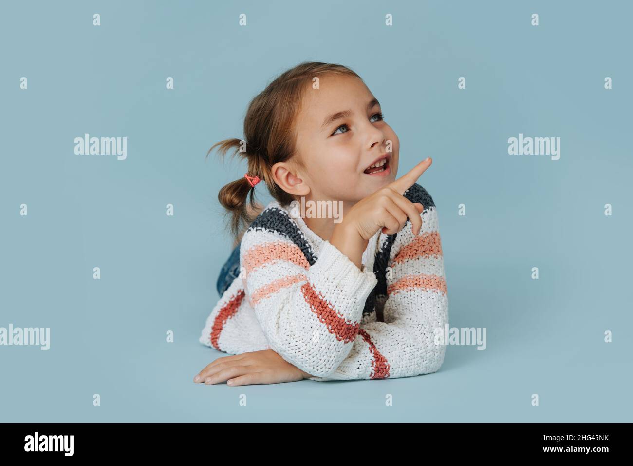 Social little girl in striped sweater lying on the floor over blue background. Talking to someone while articulating with her index finger. Stock Photo