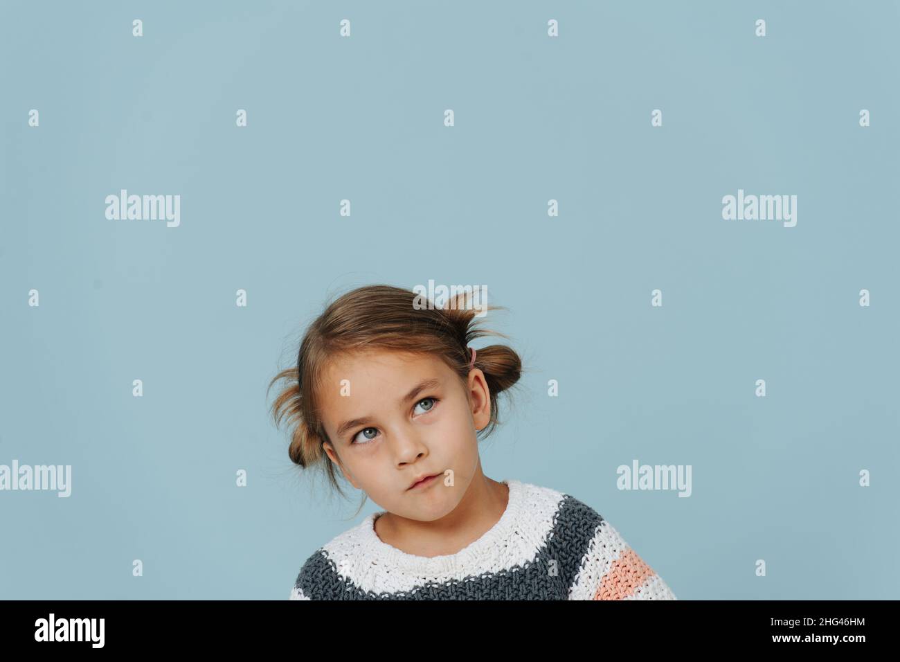Looking up tight-lipped little girl in striped sweater, hair in buns over blue. A lot of copy space around. Stock Photo