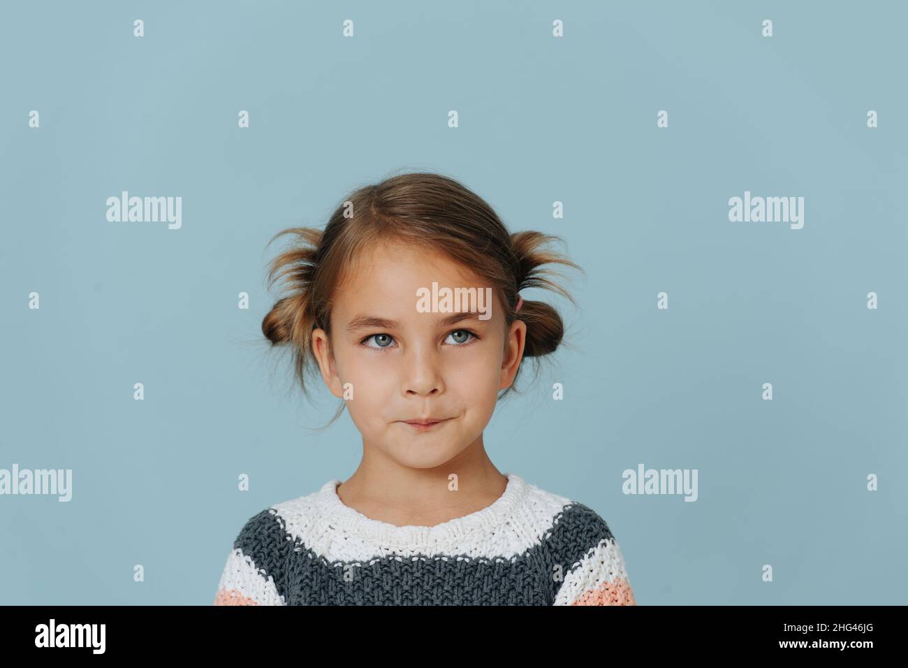 Tight-lipped little girl in striped sweater, hair in buns over blue background. Looking at someone. Stock Photo