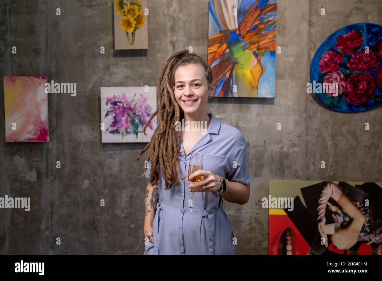 Happy young female with dreadlocks toasting with flute of champagne while standing in front of camera in art gallery Stock Photo