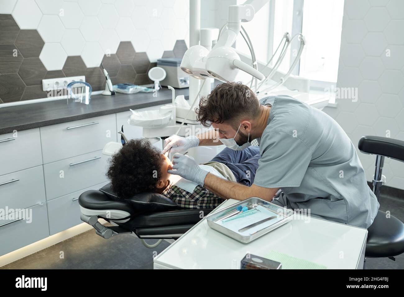 Contemporary dentist bending over little patient with cavity while drilling tooth with dental