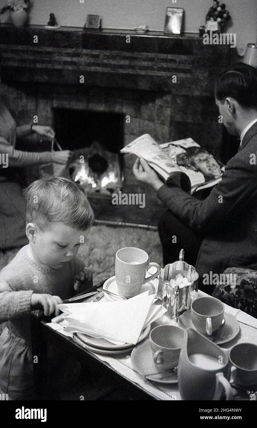 1950s, historical, tea infront of the fire, a little boy looking if there is anything cake left on the tea tray, as his father reads a magazine and his mother, by the fireplace checks the open fire, England, UK. Stock Photo