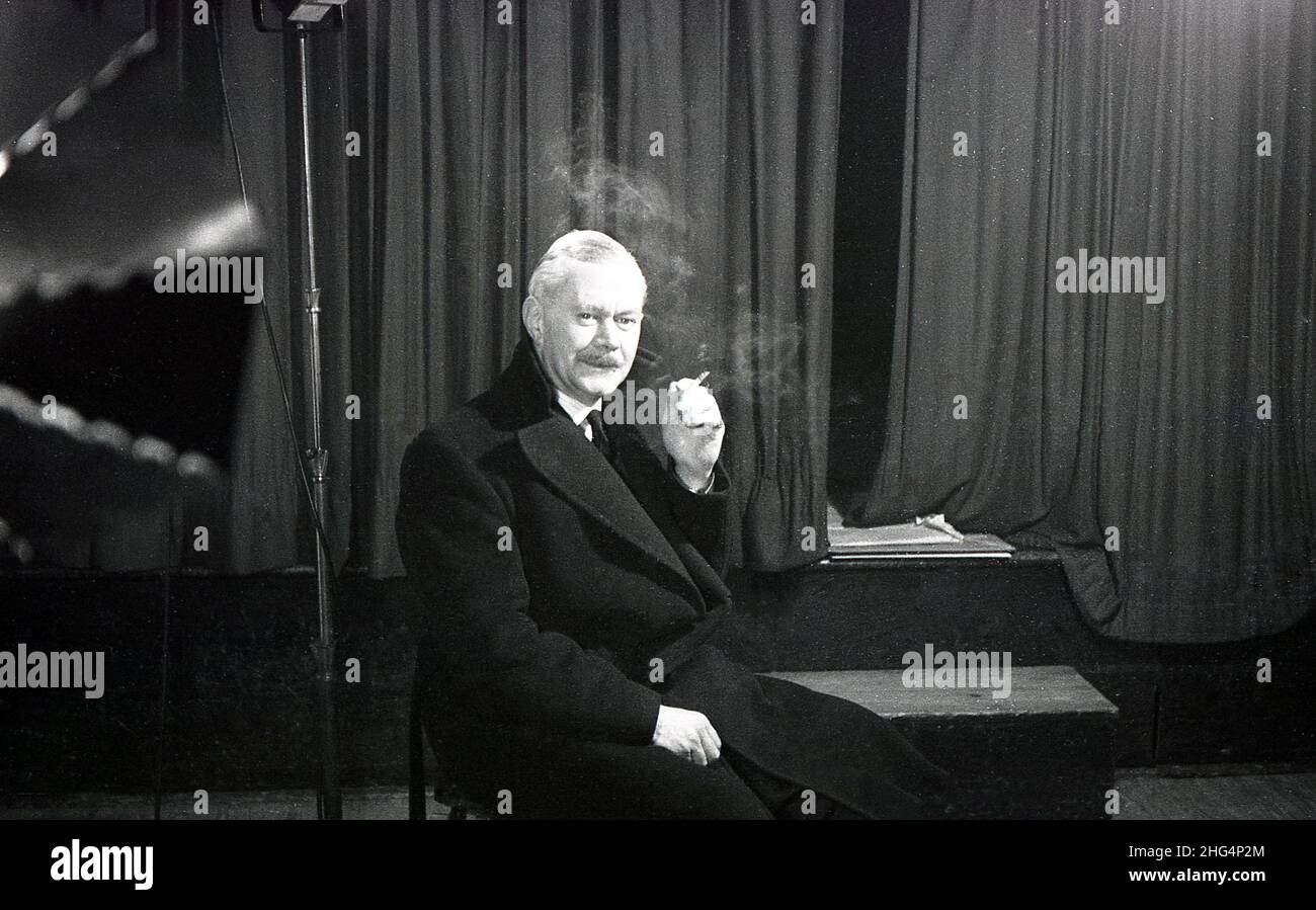 1950s, historical, a man, possibly an actor or performer in rehearsals, sitting in his overcoat by a stage and curtains on a break, having a cigarette, England, UK. Stock Photo