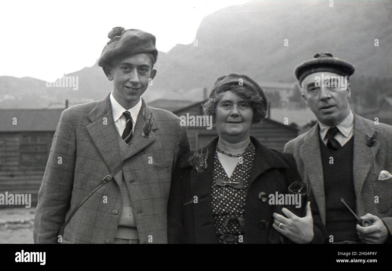 1950s, historical, a scottish family, mother, father and son standing outside near Loch Awe, Highlands, Scotland, UK. Father and son are wearing tartan jackets with ties and scottish berets. The man sports a scottish cap known as a 'Tam o'shanter', or tammie, a flat woollen bonnet, while the young man is wearing on his head, a 'Balmoral' or more formally a Balmoral bonnet, a traditional scottish hat normally worn with Highland dress. in the buttonhole or boutonniere of the jacket lapels of both males, are dried flowers, perphaps thistles, a tradition dating back to ancient  Roman times. Stock Photo