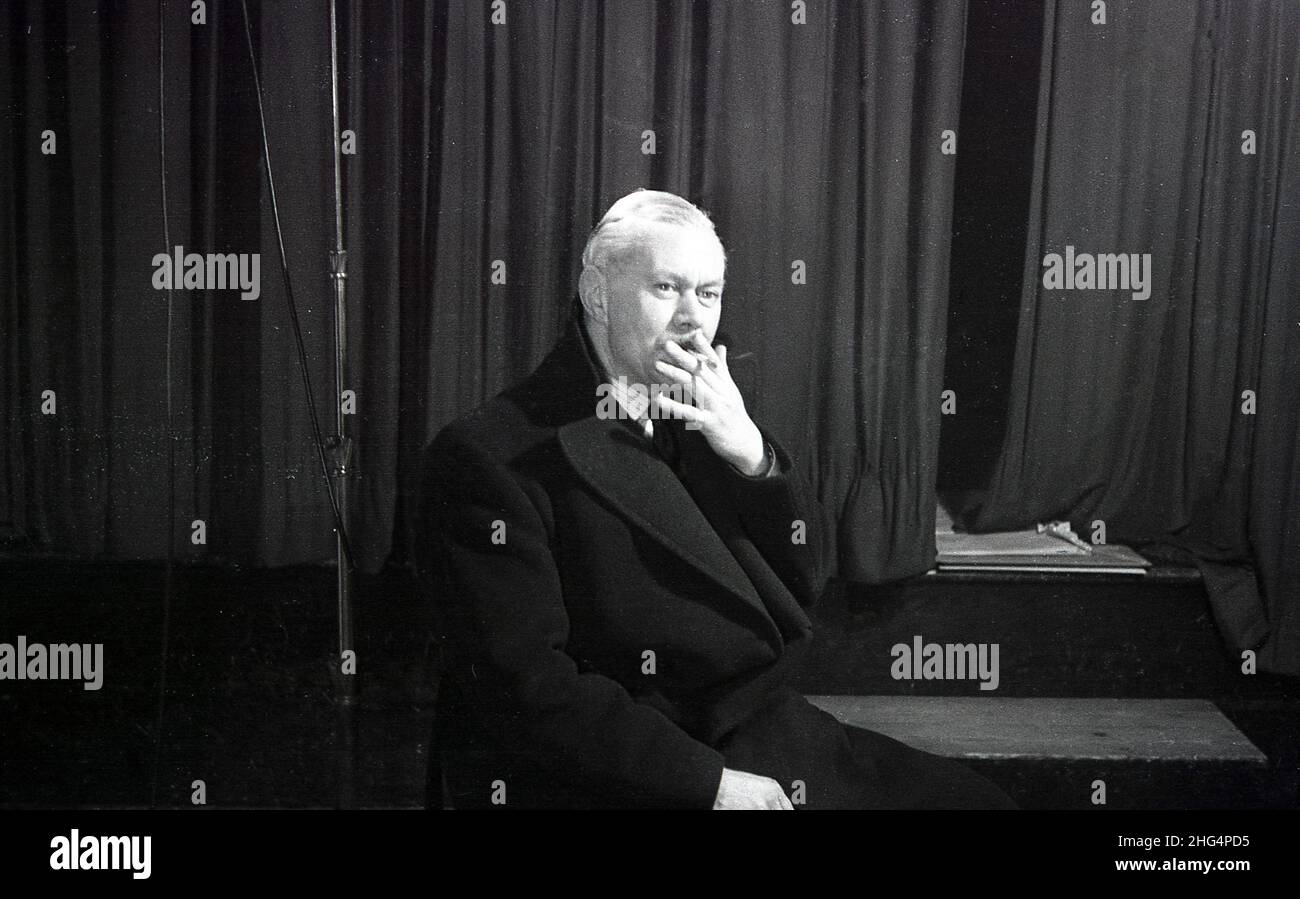 1950s, historical, a man, possibly an actor, sitting in his overcoat by a stage and curtains having a cigarette, England, UK. Stock Photo