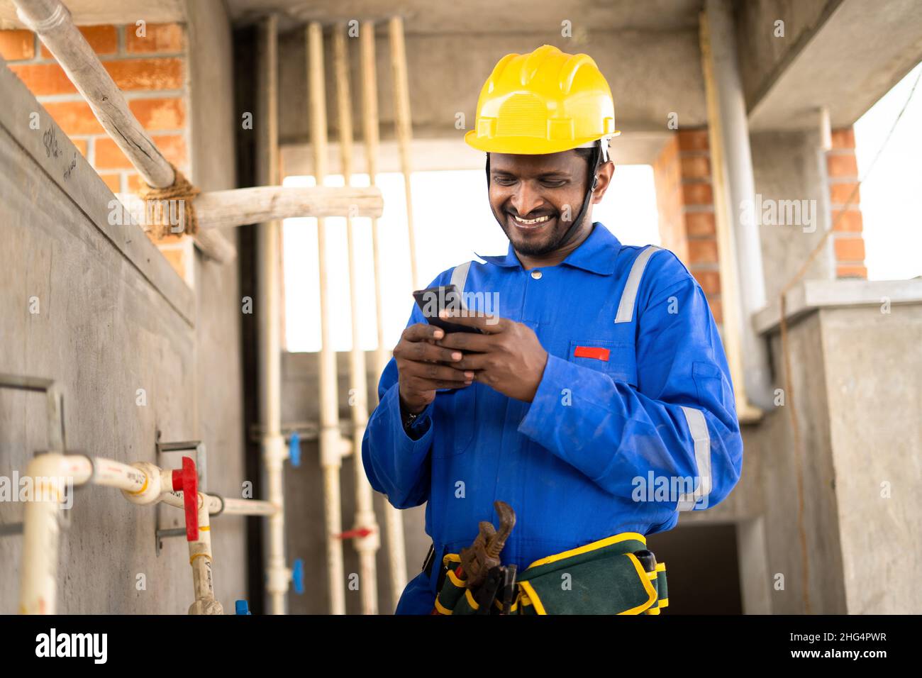 smilling young indian plumber or repairman busy by using mobile phone while working on industrial - concept of wasting of time by using social media Stock Photo