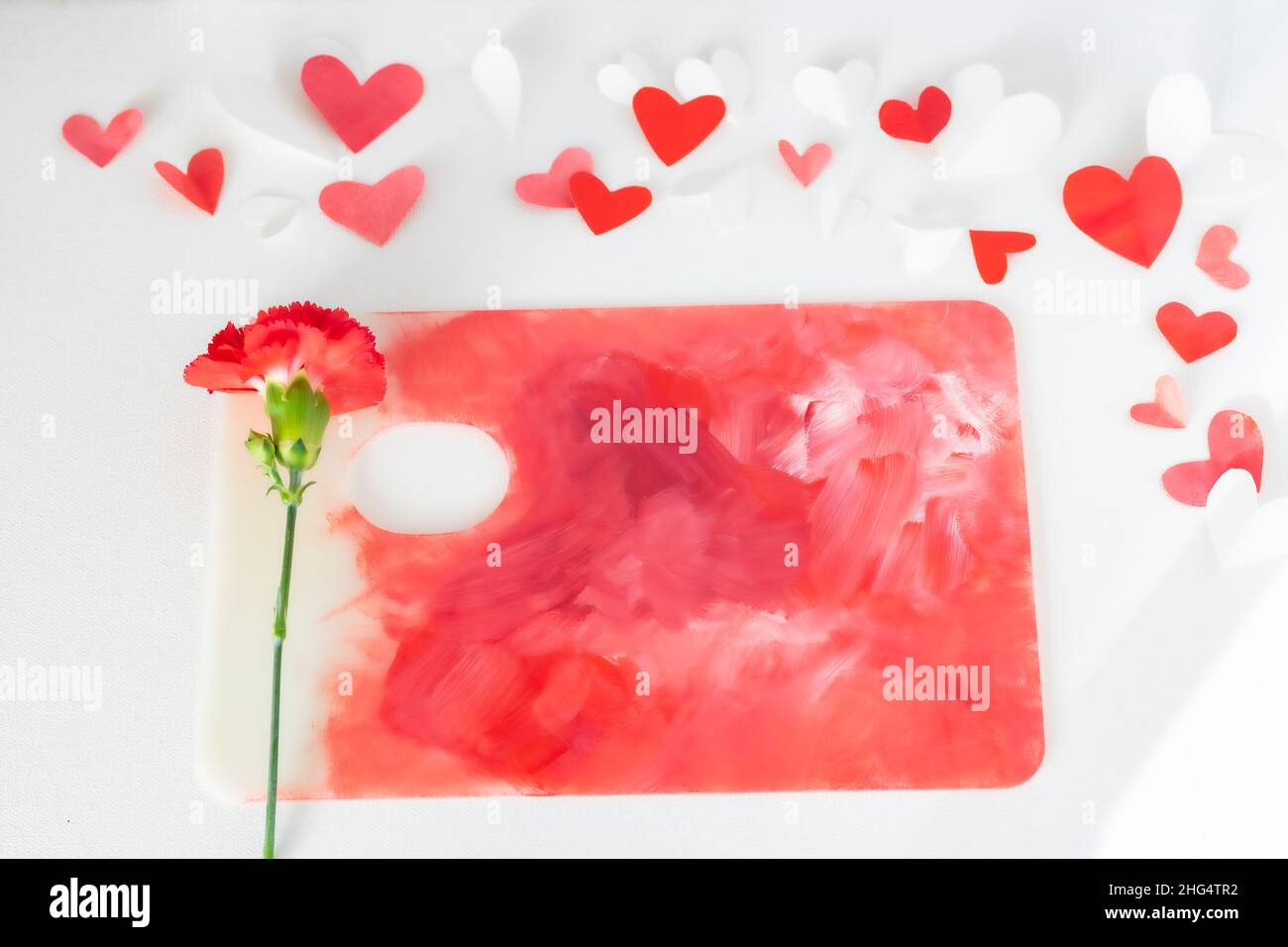 Valentines Day or wedding, invitation, scattered painted small cut out red, pink and white hearts with a red carnation on a colorful paint palette Stock Photo