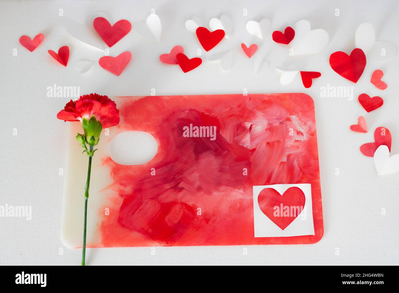 Valentines Day or wedding, invitation, scattered painted small cut out red, pink and white hearts with a red carnation on a colorful paint palette Stock Photo