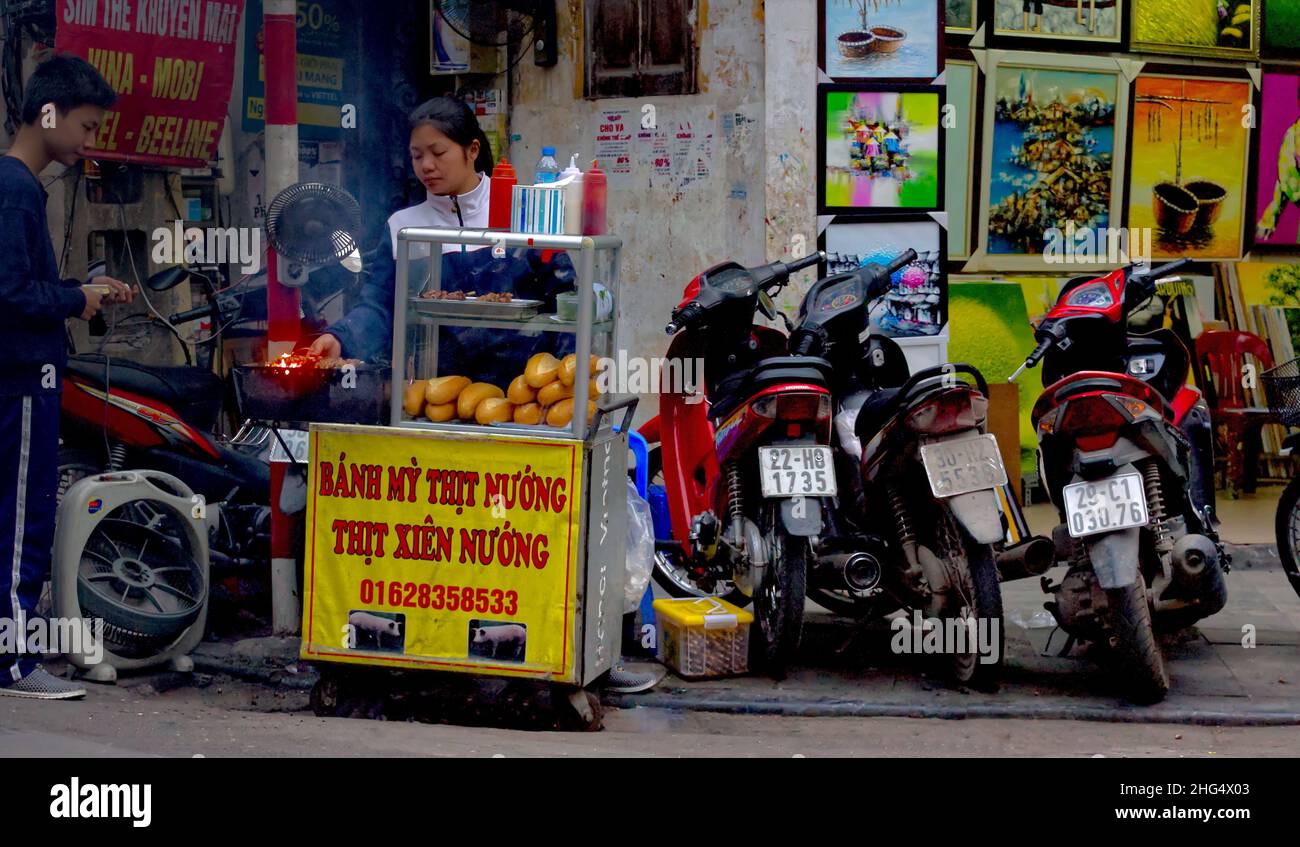 Eine junge vietnamesische Frau bietet auf einer Straßenküche Gegrilltes zum Verkauf an - ein junger Mann schaut sich das Angebot interessiert an Stock Photo