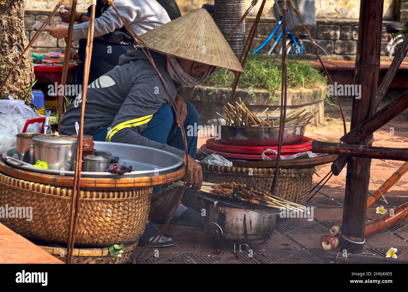Vietnamesin in gebückter Haltung mit Reis- bzw. Kegelhut grillt auf einem Markt in Hoi An Fleischspieße für den Straßenverkauf Stock Photo