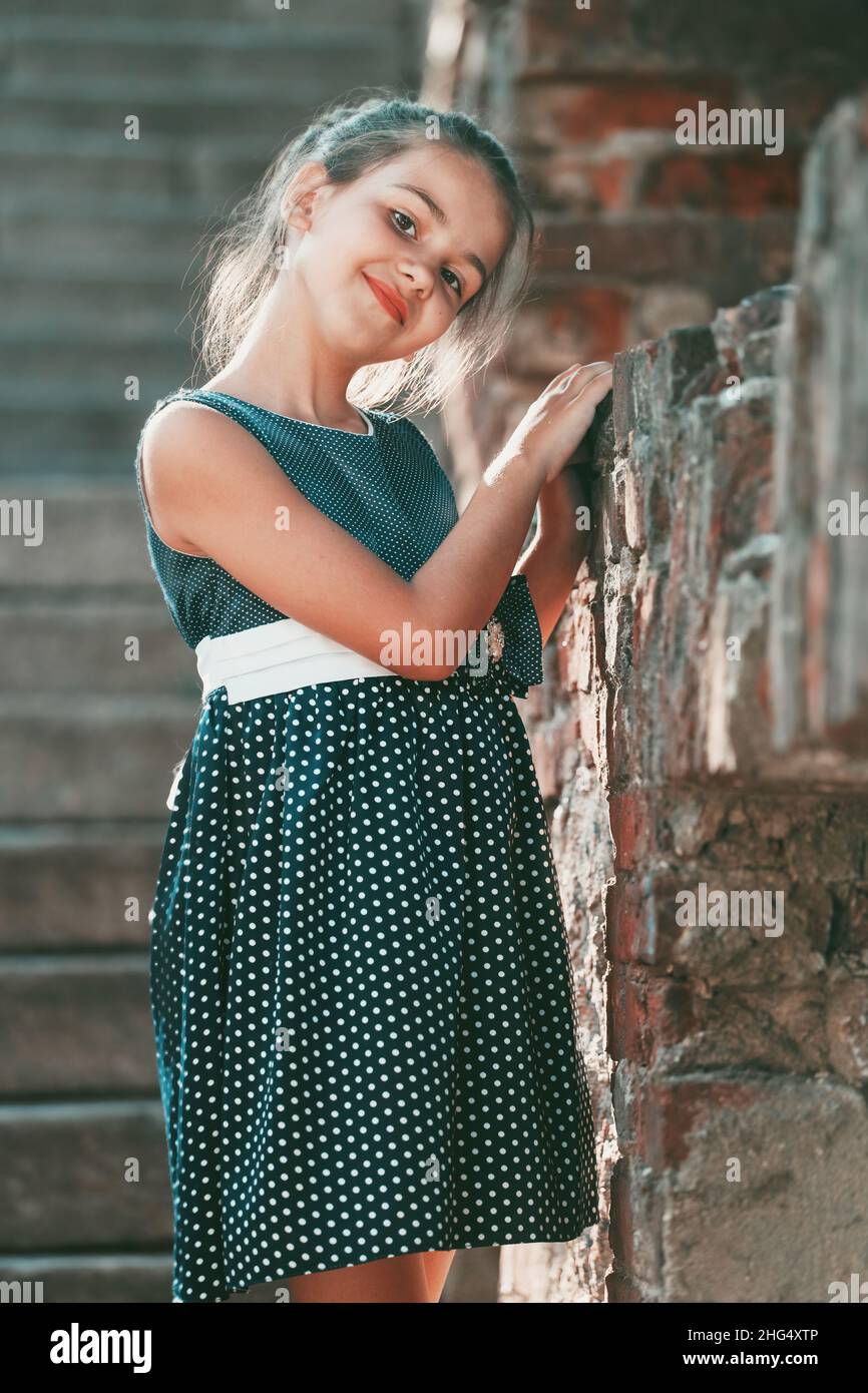 a young brunette girl in a blue polka dot dress stands against the wall ...