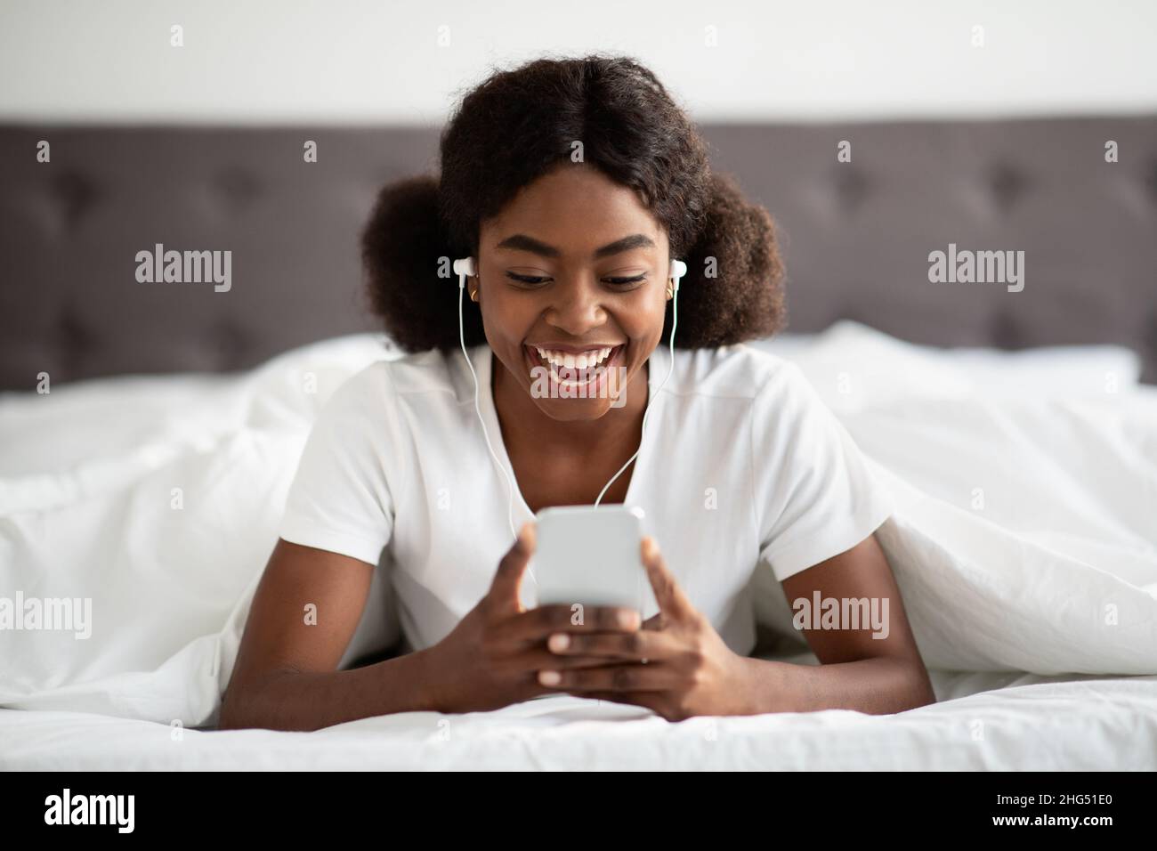 Happy young black woman laying in bed with smartphone, earphones Stock ...