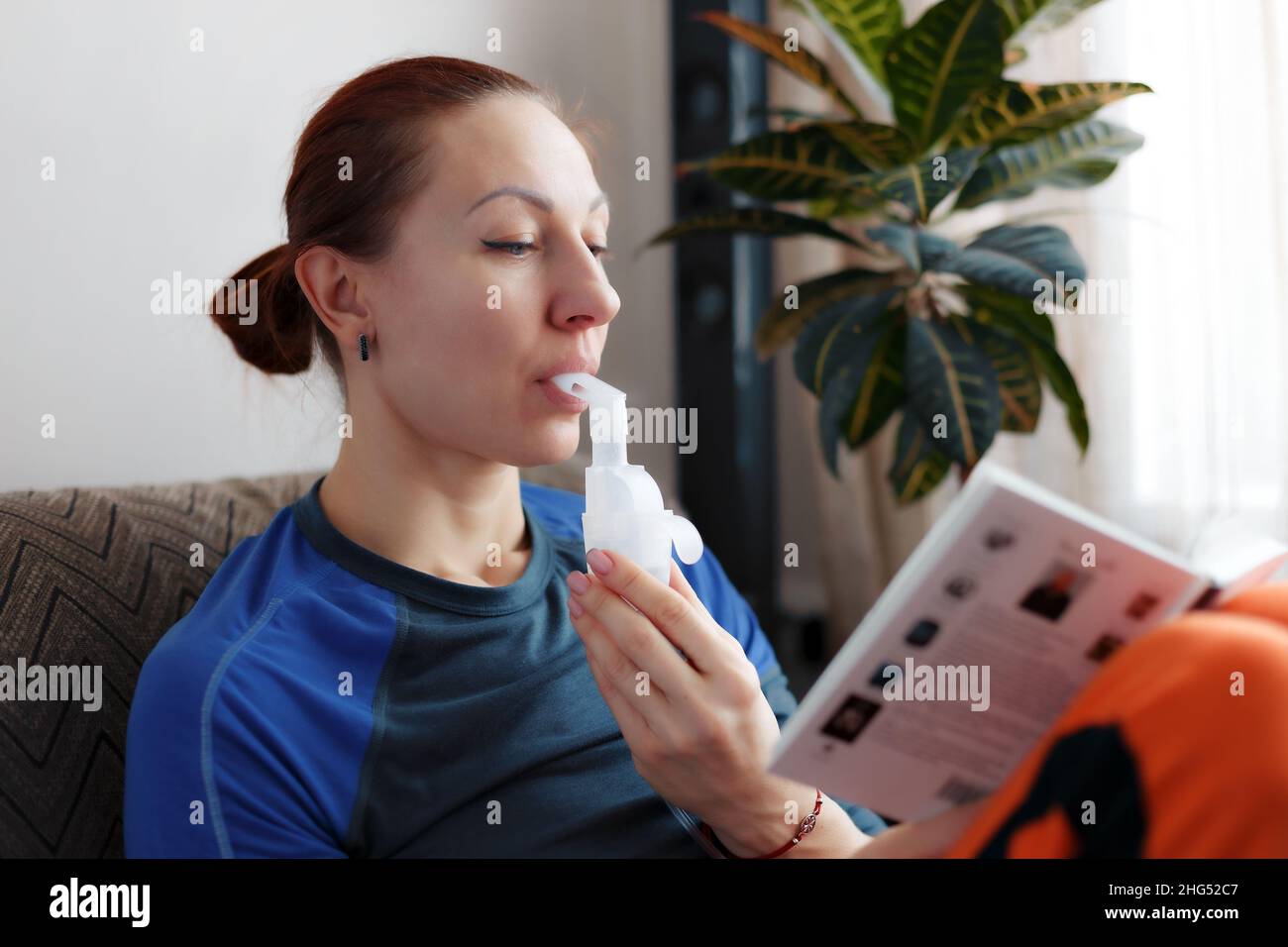 A caucasian woman is reading a book and holding a nebulizer tube, inhaling vapors of medicines into her lungs. Treating asthma or cold flu at home. Inhalation in respiratory diseases. Stock Photo