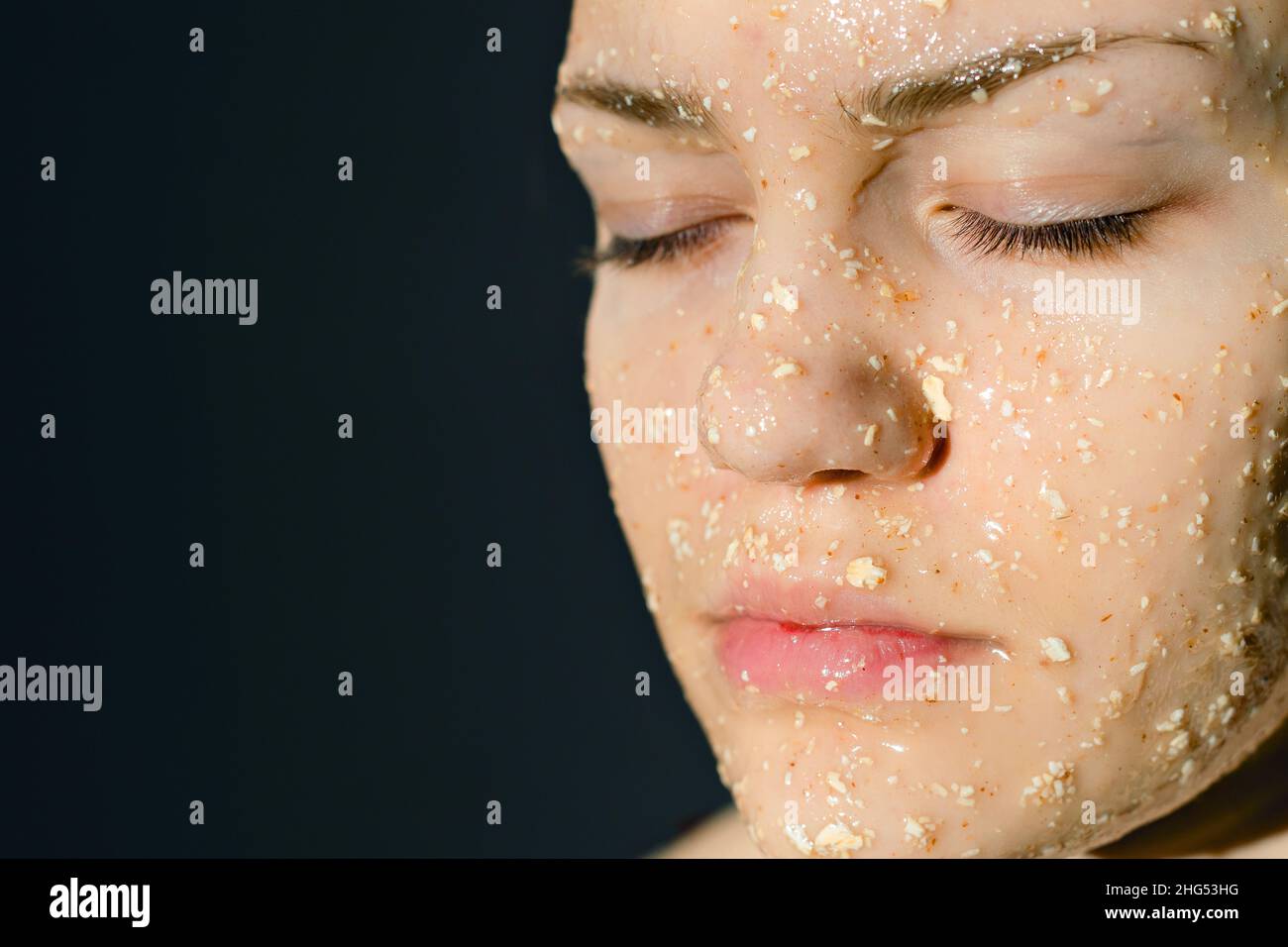 Face of girl with cosmetic mask of oatmeal with honey. Prevention of acne in adolescents. Woman's face with her eyes closed on dark background. Stock Photo
