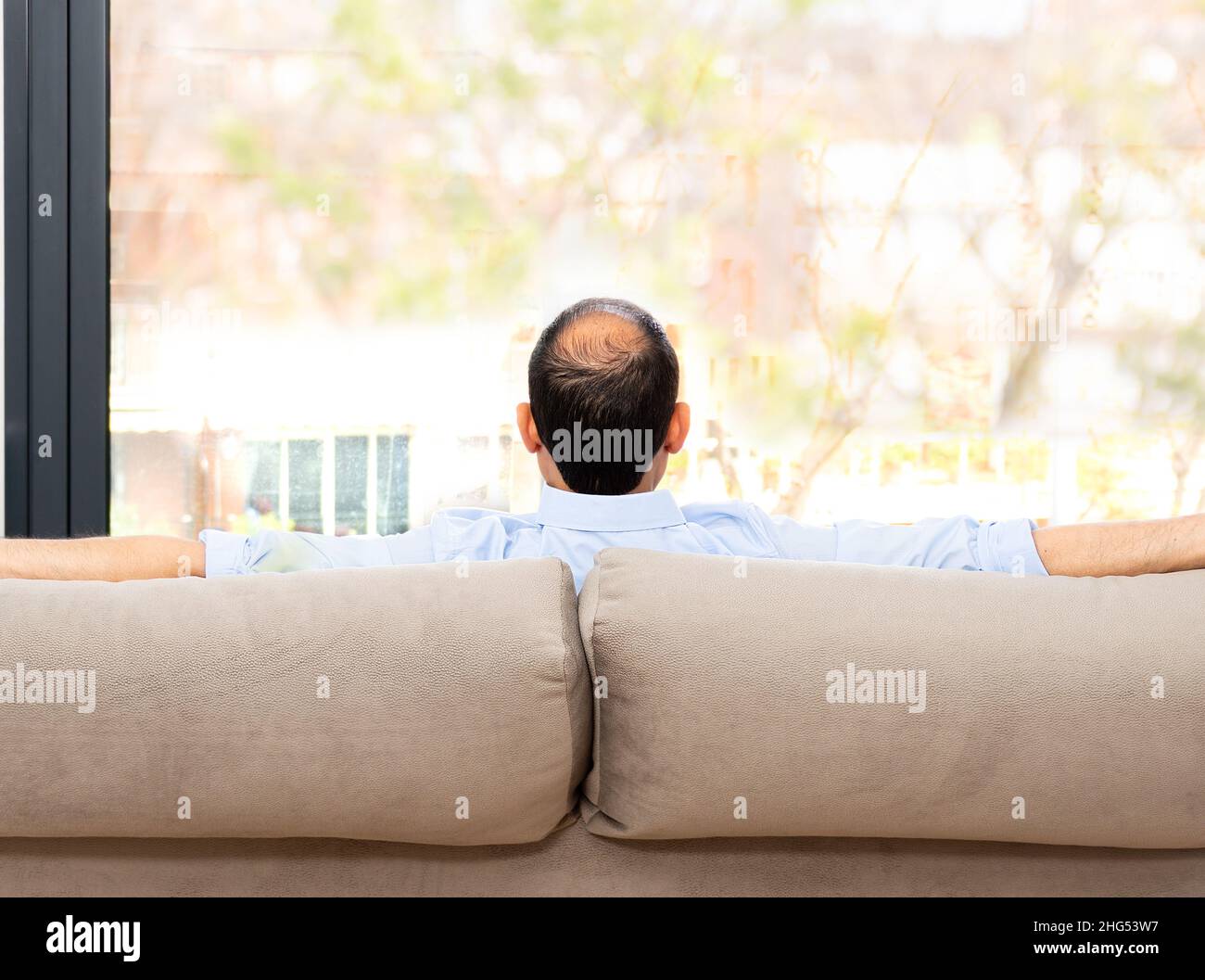 Rear view of a bald and carefree man sitting on a sofa at home and looking outdoors through the window at home Stock Photo