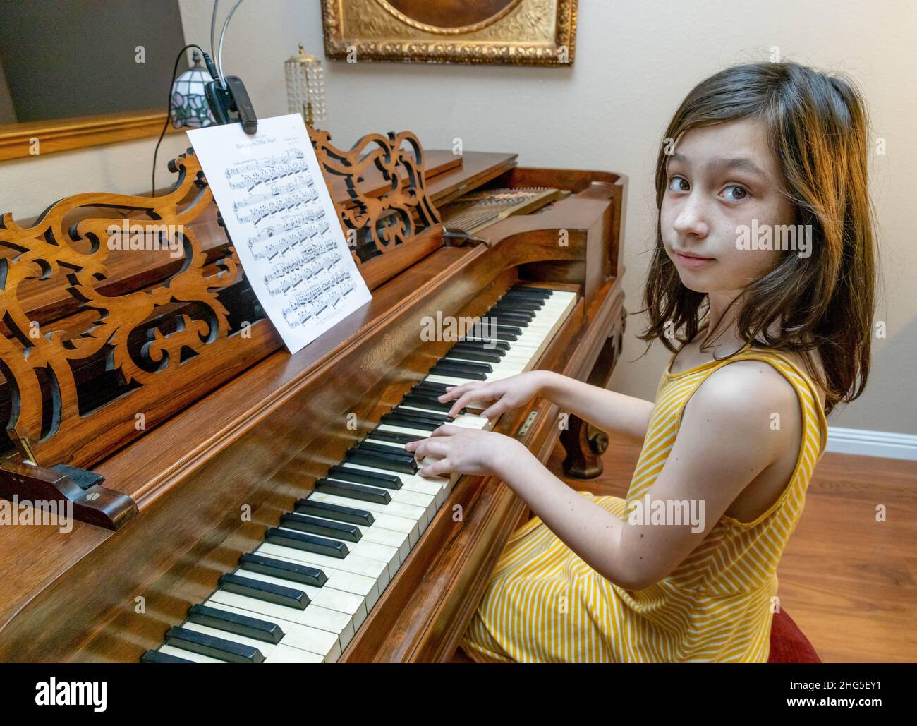 A young girl in a yellow dress playing a rare antique Chickering & Sons Square Grand piano built in 1867 with an antique painting in a gold frame. Stock Photo