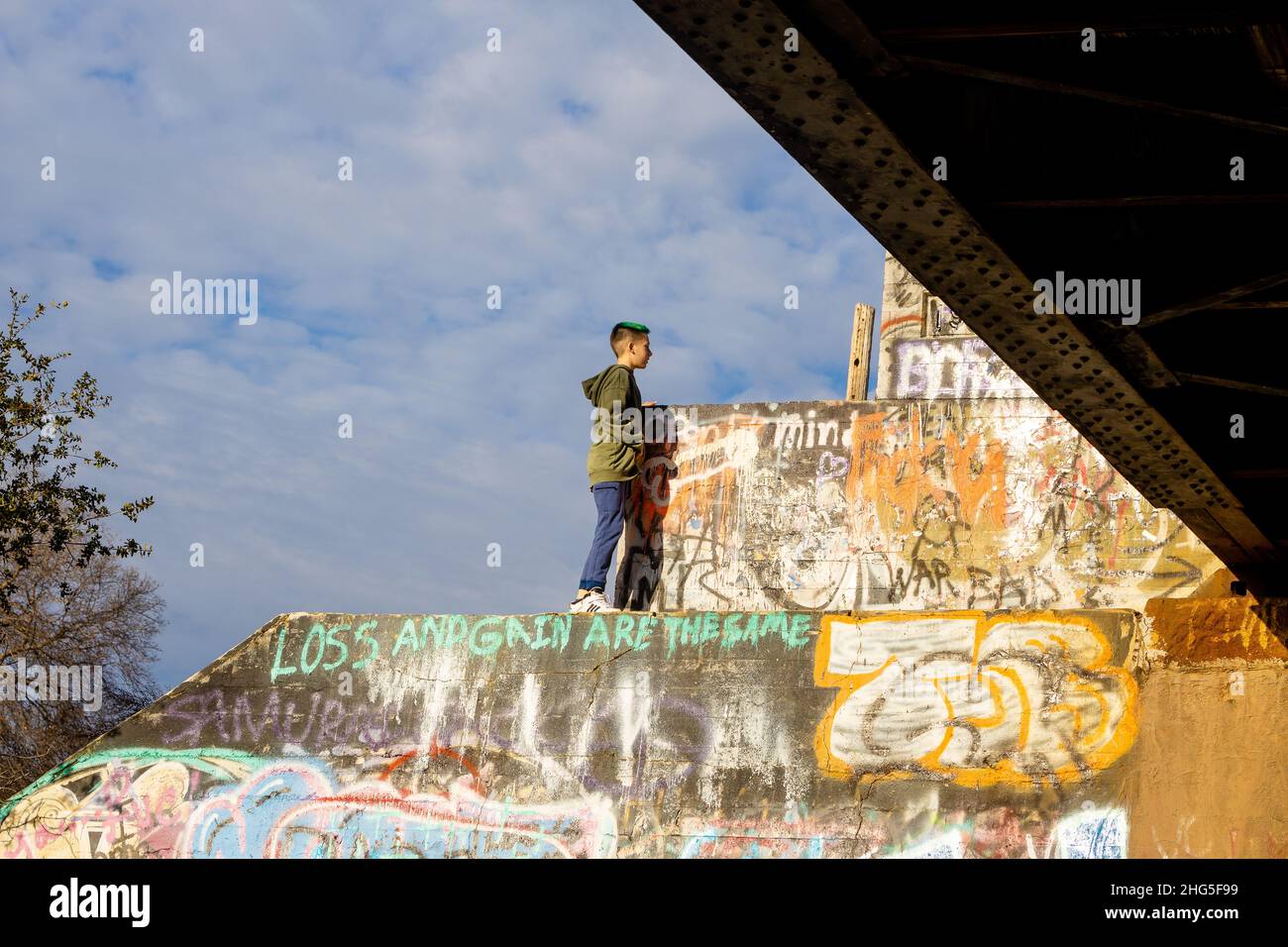 Boy on top of railroad pillars platforms next to a train bridge with graffiti and blue sky and clouds Stock Photo