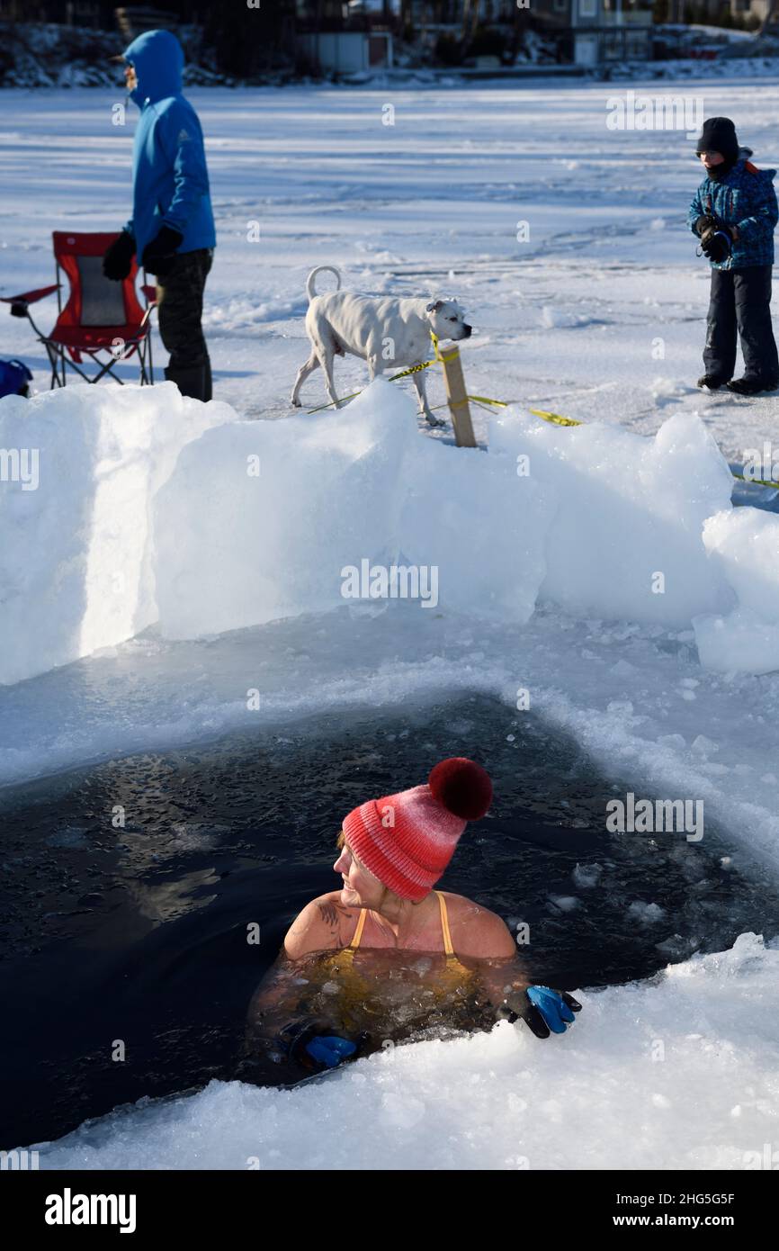Woman with hat in ice hole for a cold water swim in winter with dog and spectators at Kempenfelt Bay Lake Simcoe Barrie Ontario Stock Photo