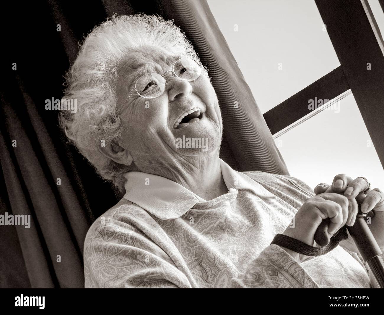 ELDERLY LADY Laughing happy independent senior old age elderly woman sitting by window with walking stick inside her light modern home B&W treatment Stock Photo