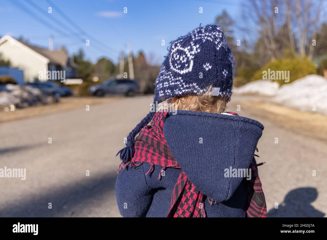 Back view of infant in winter clothing with thick coat, and blue wooly hat on a cold sunny day with blurred street view in background and copy space. Stock Photo