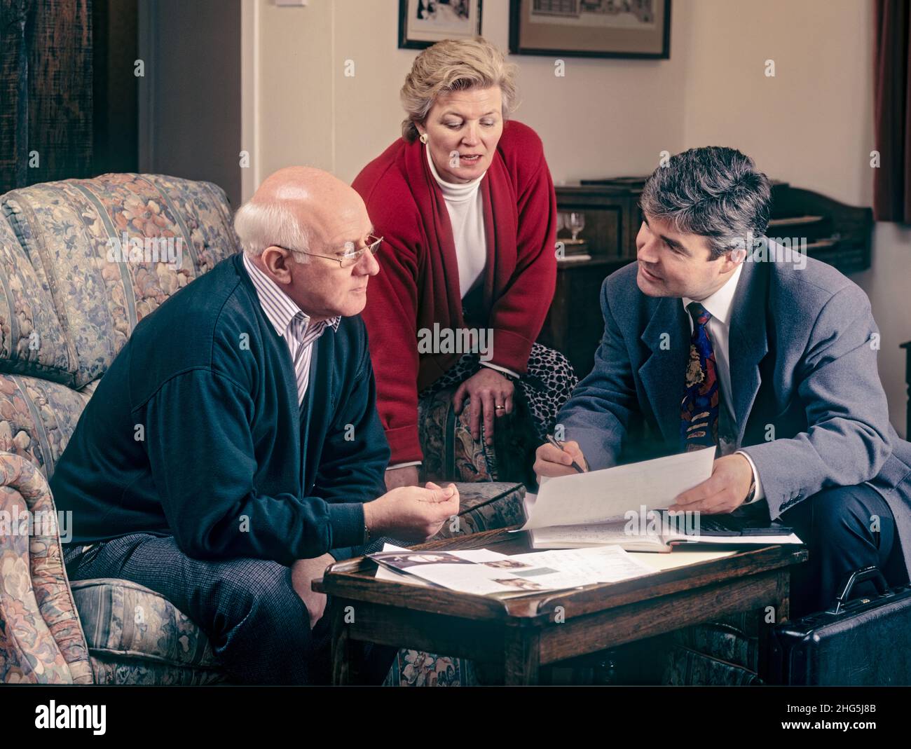 1990s Financial advisor with mature retired elderly couple at home 1990s era, talking to salesman sales person man, with variety of paperwork on table. Financial planning, home improvements, legal services, will wills writing etc. Archive 1990's home visit services salesman Stock Photo