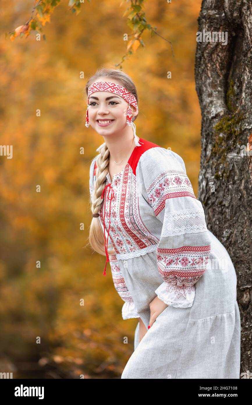 Young attractive woman smiles in folk traditional slavic dress near tree at nature in autumn forest. Portrait of beautiful girl in ethnic clothes Stock Photo