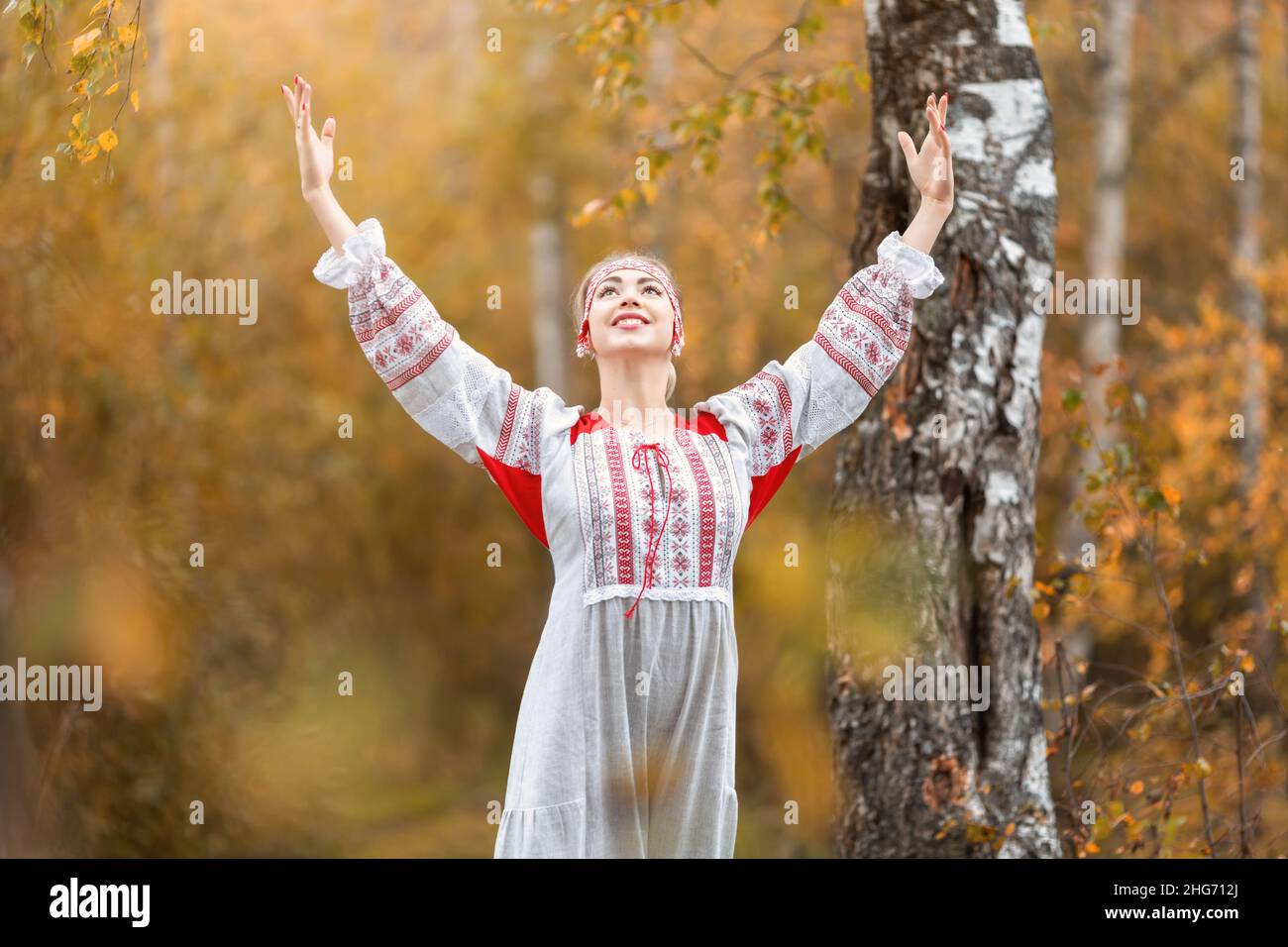 Portrait of young beautiful smiling woman in folk traditional slavic clothes waving hands  and asking for blessing from spirits at nature in autumn fo Stock Photo