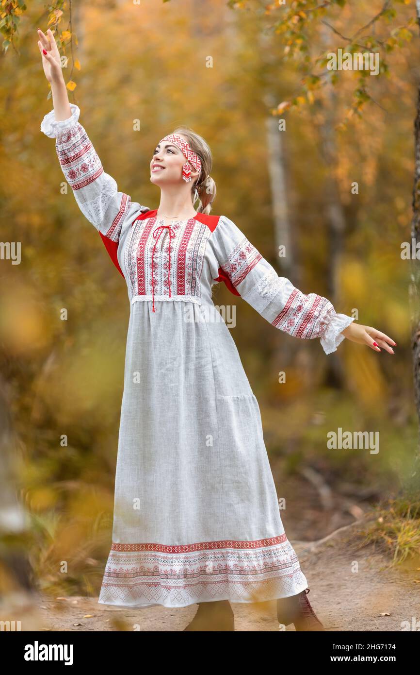 Portrait of young beautiful smiling woman in folk traditional slavic clothes greets nature spirits by hand in autumn forest. Stock Photo