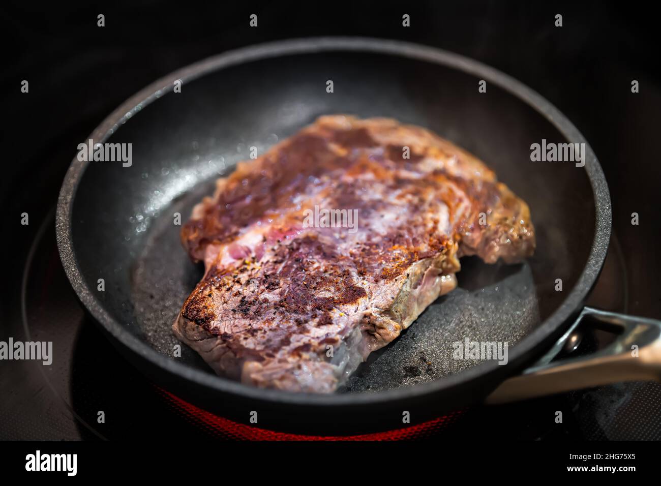 Closeup of brown charred crust on rare grass-fed ribeye rib eye meat steak cooking on non-stick frying pan grill with pepper salt seasoning Stock Photo