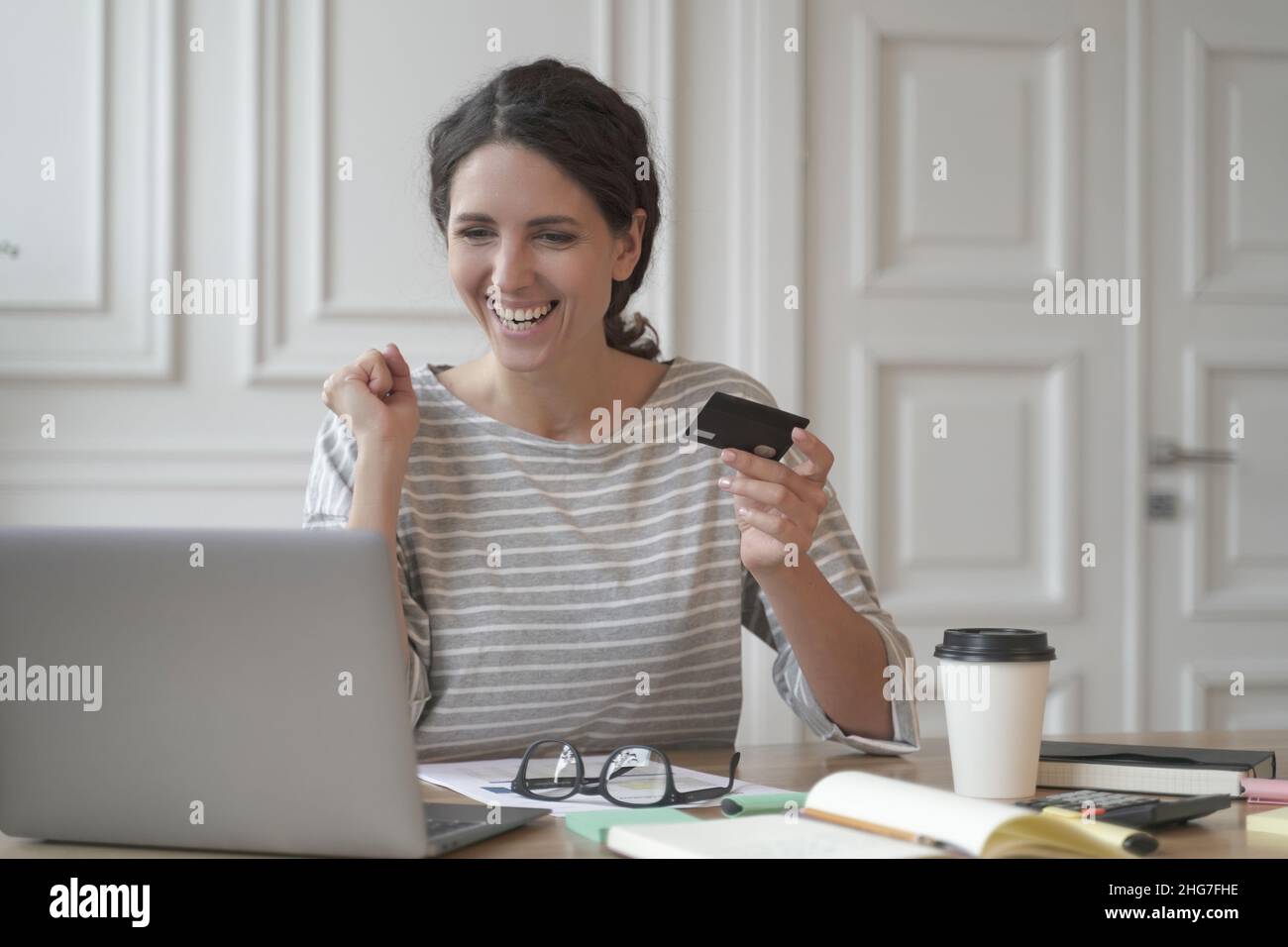 Young happy Spanish woman hold credit card and feeling excited while doing online shopping on laptop Stock Photo