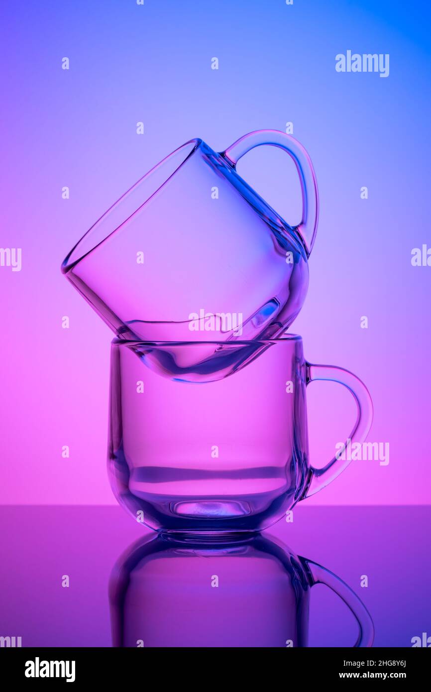 Two empty tea cups on a pink and blue neon background. Drinking glass. Dark silhouette, night illuminated effect. Glassware. Transparent tableware, di Stock Photo