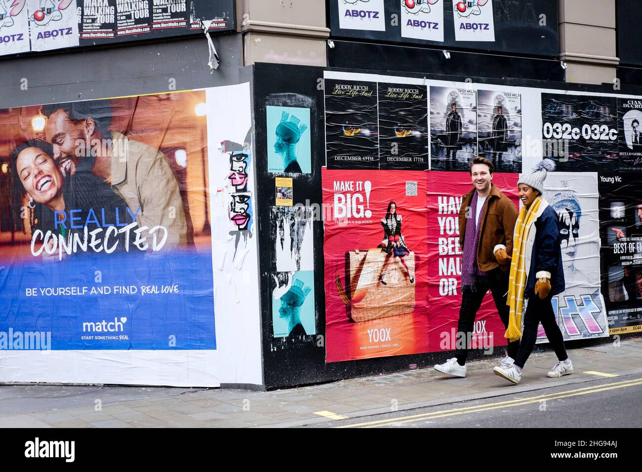 Smiling, young couple walk past wall covered with fly posters, one advertising a dating website. London, UK. Stock Photo