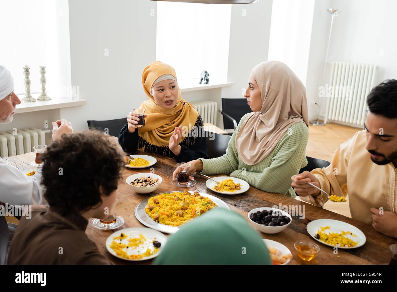 middle aged asian woman pointing with hand while talking to multiethnic muslim family during dinner Stock Photo