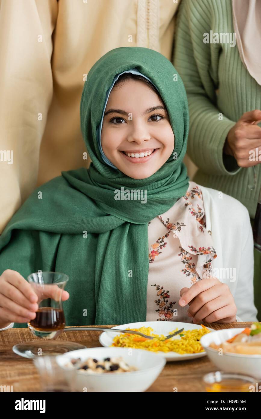 happy muslim girl in hijab looking at camera while holding glass of tea ...